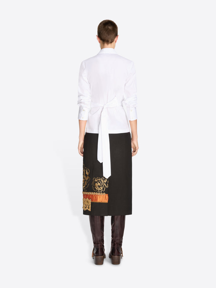 Back view of a model standing against a light gray background wearing a white long-sleeve shirt, a black midi skirt with ornate gold and printed motifs and dark brown knee-high boots.