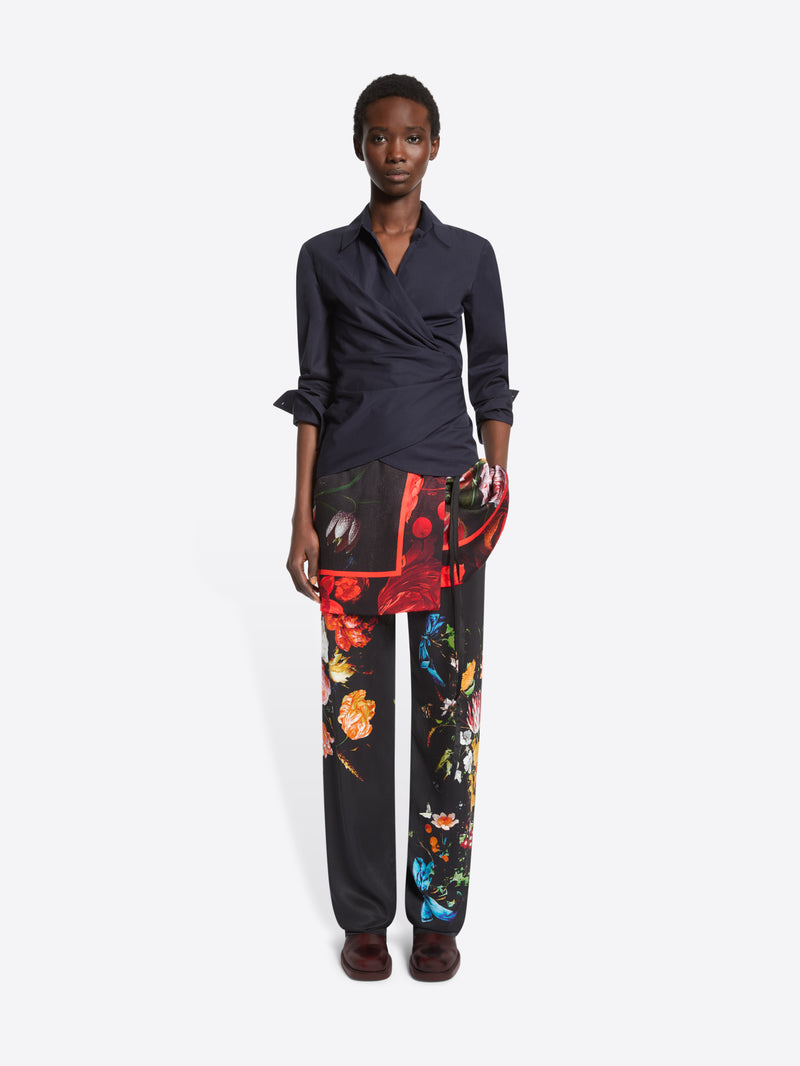 A model stands against a light gray studio background wearing a dark navy wrap-style shirt, black wide-leg trousers with a vivid multicolor floral print, printed skirt and dark brown shoes.