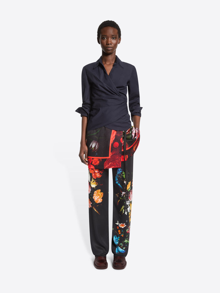 A model stands against a light gray studio background wearing a dark navy wrap-style shirt, black wide-leg trousers with a vivid multicolor floral print, printed skirt and dark brown shoes.