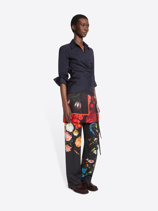 A model stands against a light gray studio background wearing a dark navy wrap-style shirt, black wide-leg trousers with a vivid multicolor floral print, printed skirt and dark brown shoes.