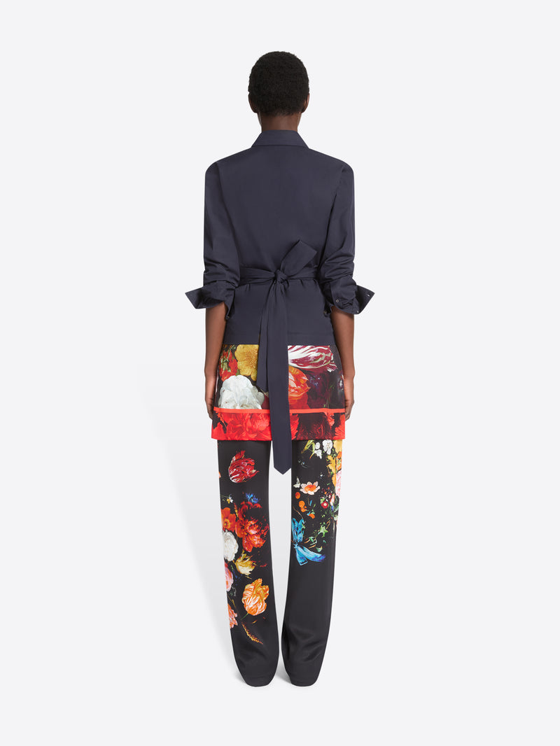 Back view of a model standing against a light gray studio background wearing a dark navy wrap-style shirt, black wide-leg trousers with a vivid multicolor floral print, printed skirt and dark brown shoes.