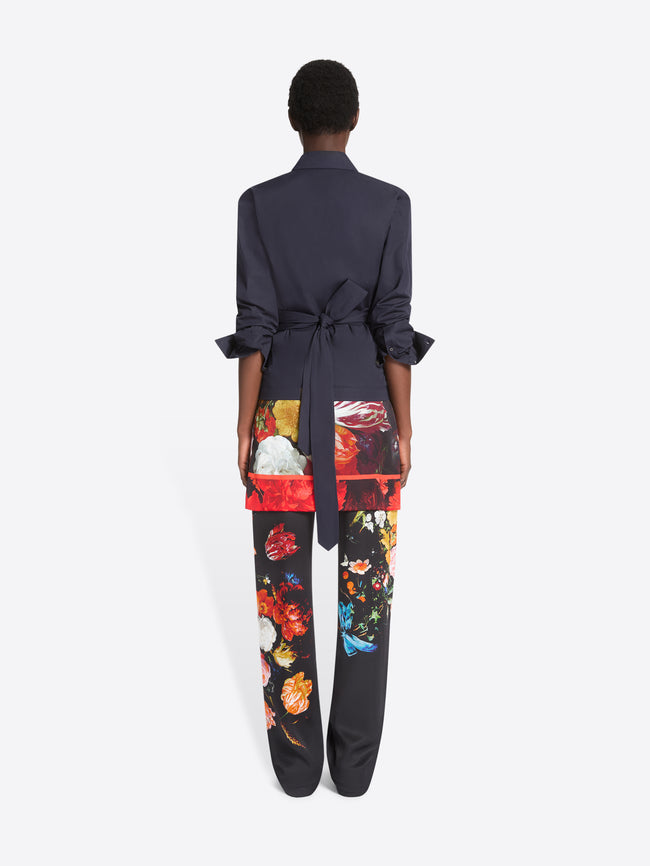 Back view of a model standing against a light gray studio background wearing a dark navy wrap-style shirt, black wide-leg trousers with a vivid multicolor floral print, printed skirt and dark brown shoes.