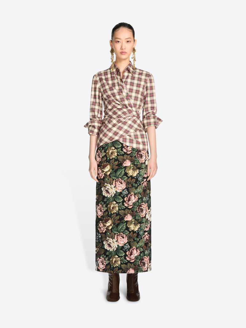 A model stands against a light gray background wearing a brown and beige plaid wrap shirt with ruffled sleeves and a dark green floral midi skirt, paired with dark brown ankle boots.