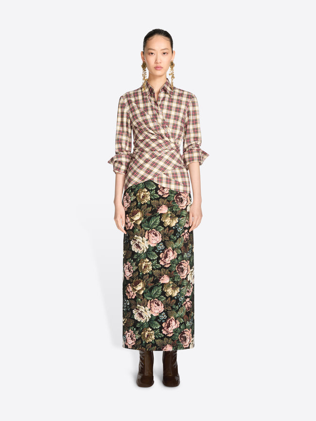 A model stands against a light gray background wearing a brown and beige plaid wrap shirt with ruffled sleeves and a dark green floral midi skirt, paired with dark brown ankle boots.