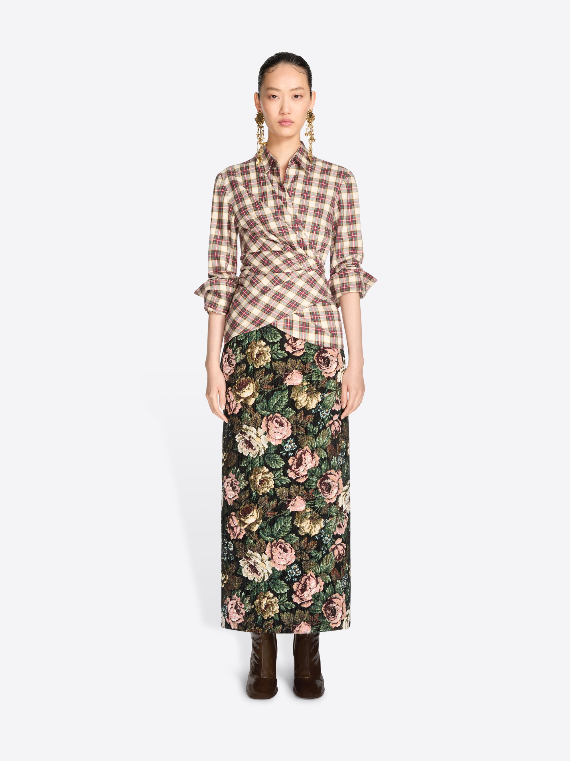 A model stands against a light gray background wearing a brown and beige plaid wrap shirt with ruffled sleeves and a dark green floral midi skirt, paired with dark brown ankle boots.