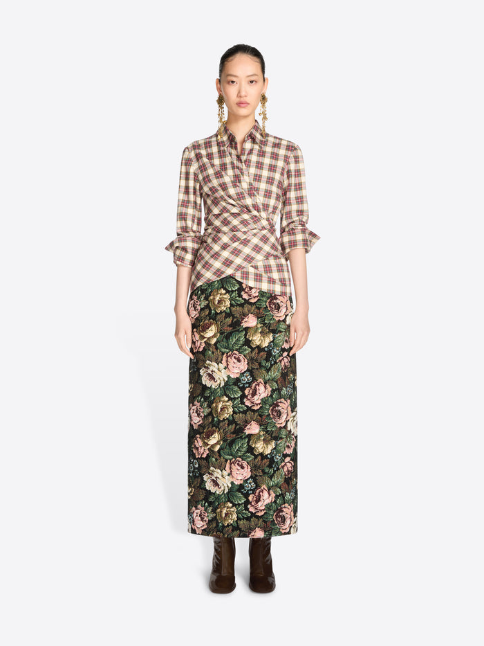 A model stands against a light gray background wearing a brown and beige plaid wrap shirt with ruffled sleeves and a dark green floral midi skirt, paired with dark brown ankle boots.