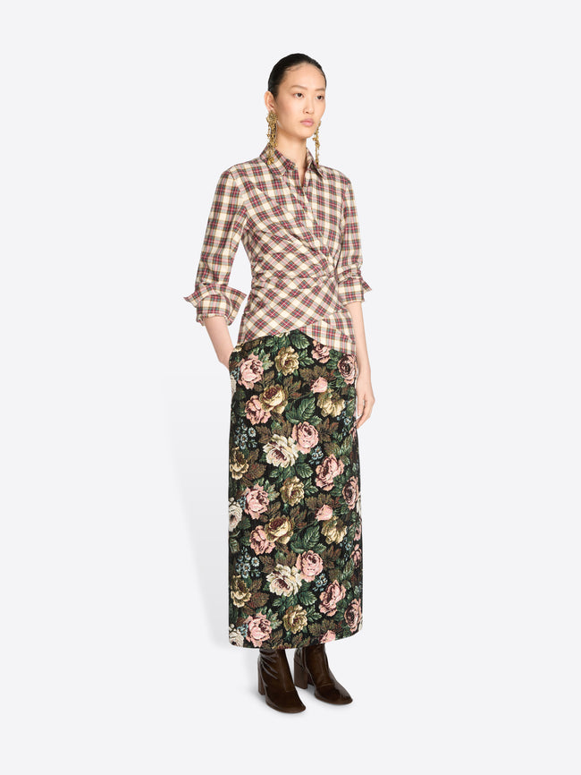 A model stands against a light gray background wearing a brown and beige plaid wrap shirt with ruffled sleeves and a dark green floral midi skirt, paired with dark brown ankle boots.
