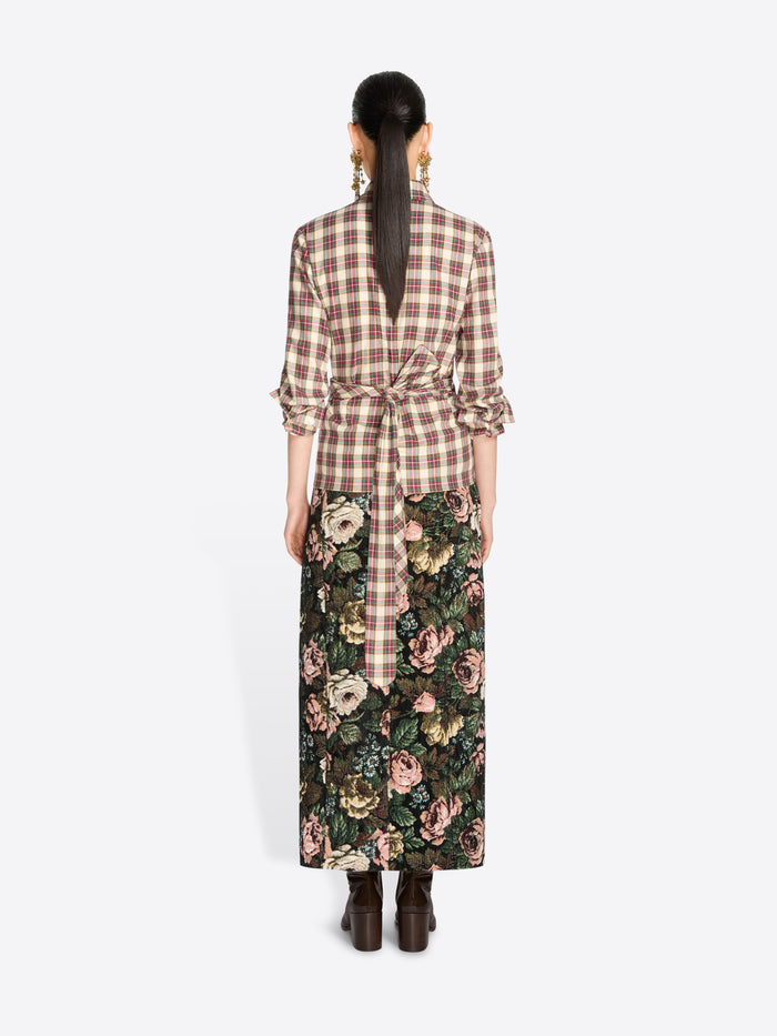 Back view of a model standing against a light gray background wearing a brown and beige plaid wrap shirt with ruffled sleeves and a dark green floral midi skirt, paired with dark brown ankle boots.