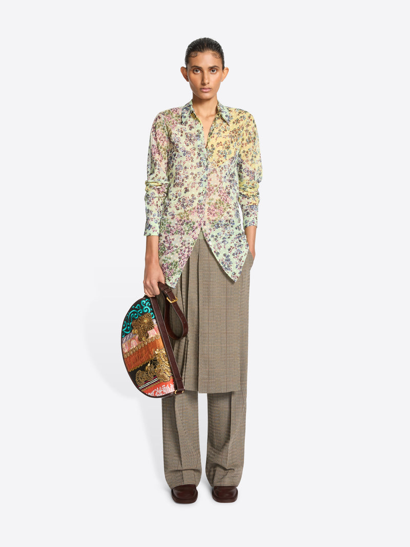 A model stands against a light gray background wearing a long-sleeve button-up shirt with a dense multicolor floral print, paired with loose taupe wide-leg pants and skirt on top, holding a small oval handbag with a colorful abstract print.