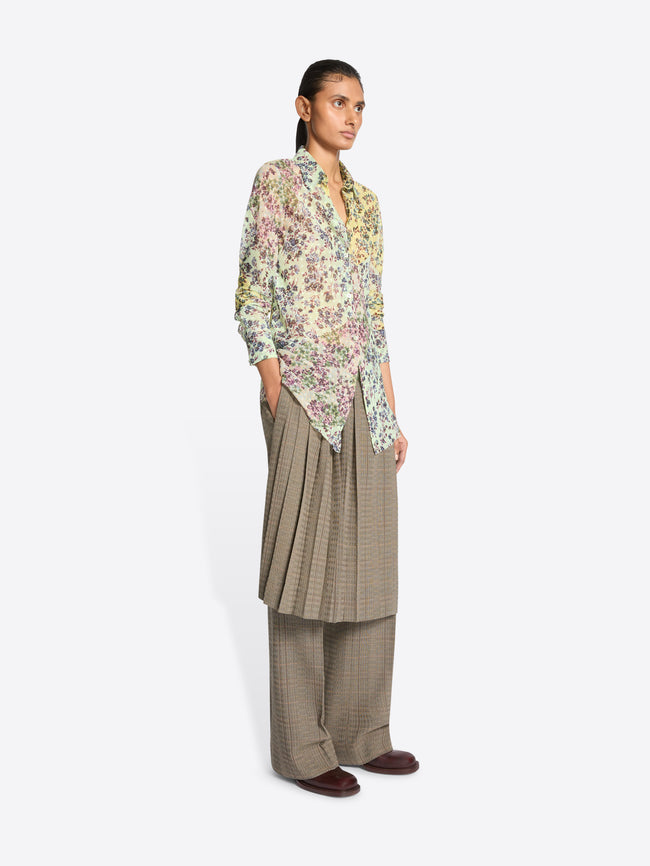 A model stands against a light gray background wearing a long-sleeve button-up shirt with a dense multicolor floral print, paired with loose taupe wide-leg pants and skirt on top.