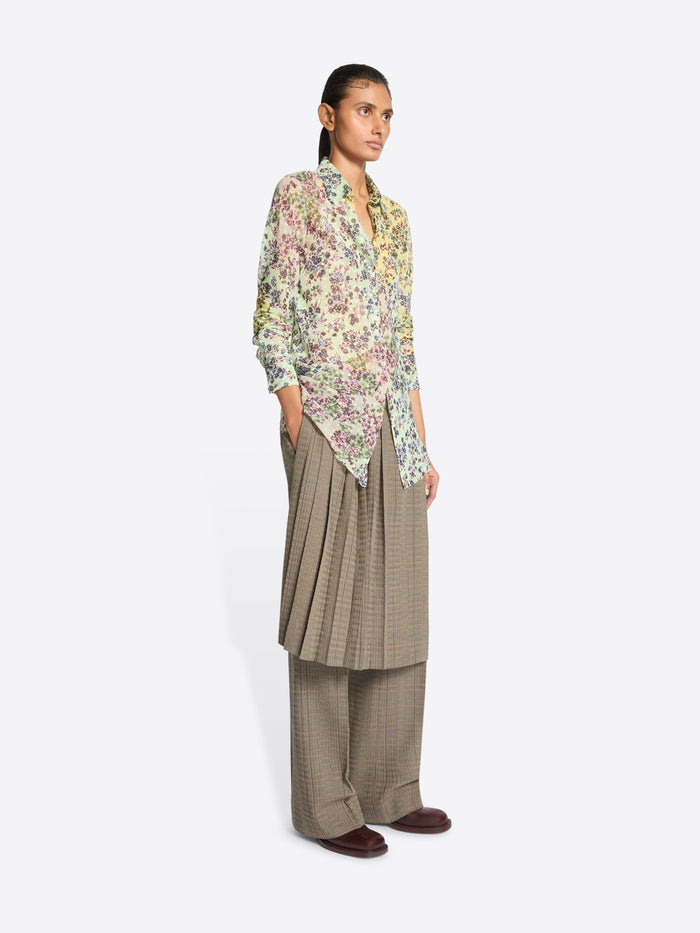 A model stands against a light gray background wearing a long-sleeve button-up shirt with a dense multicolor floral print, paired with loose taupe wide-leg pants and skirt on top.