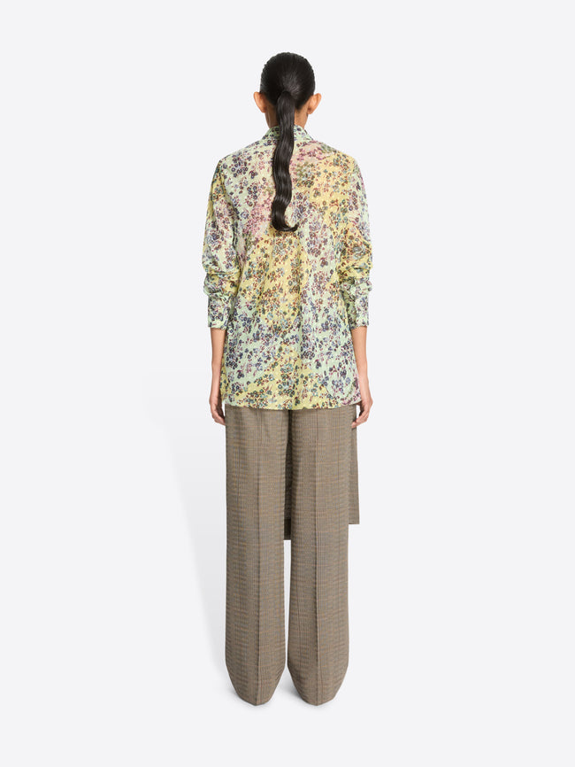 Back view of a model standing against a light gray background wearing a long-sleeve button-up shirt with a dense multicolor floral print, paired with loose taupe wide-leg pants and skirt on top.
