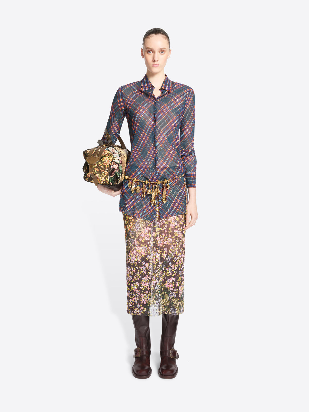 A model stands against a light gray background wearing a long-sleeve collared shirt with a muted plaid pattern in blue, red, and gray, paired with a floral midi skirt and dark brown knee-high boots, holding a small patterned handbag.