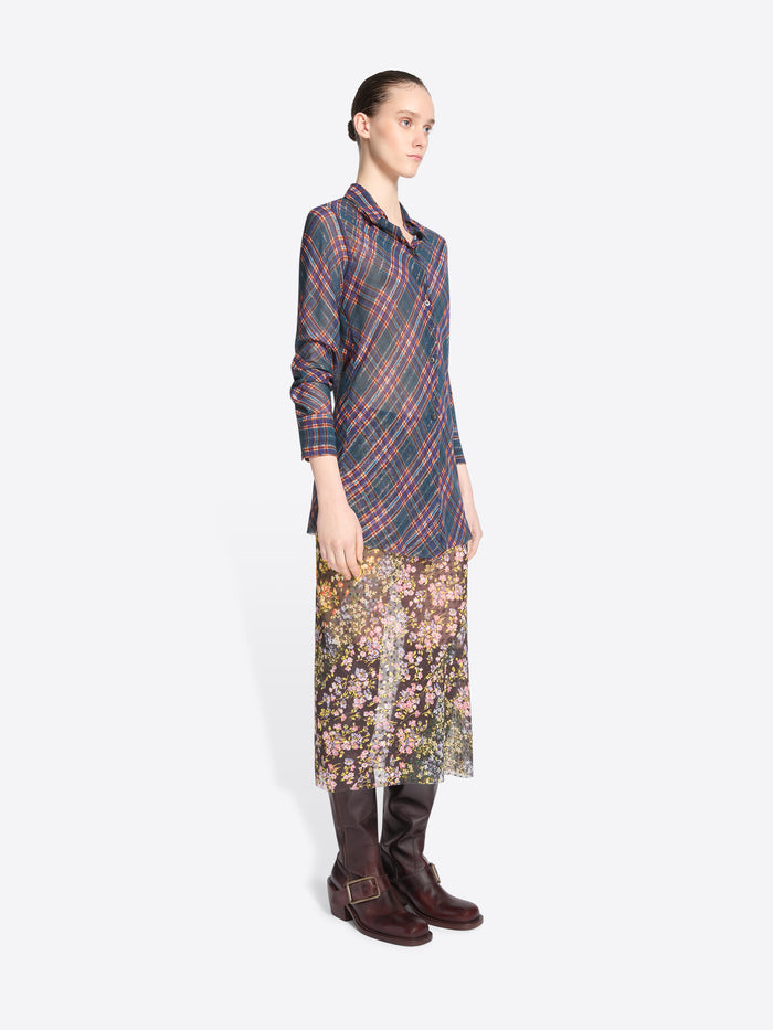 A model stands against a light gray background wearing a long-sleeve collared shirt with a muted plaid pattern in blue, red, and gray, paired with a floral midi skirt and dark brown knee-high boots. 
