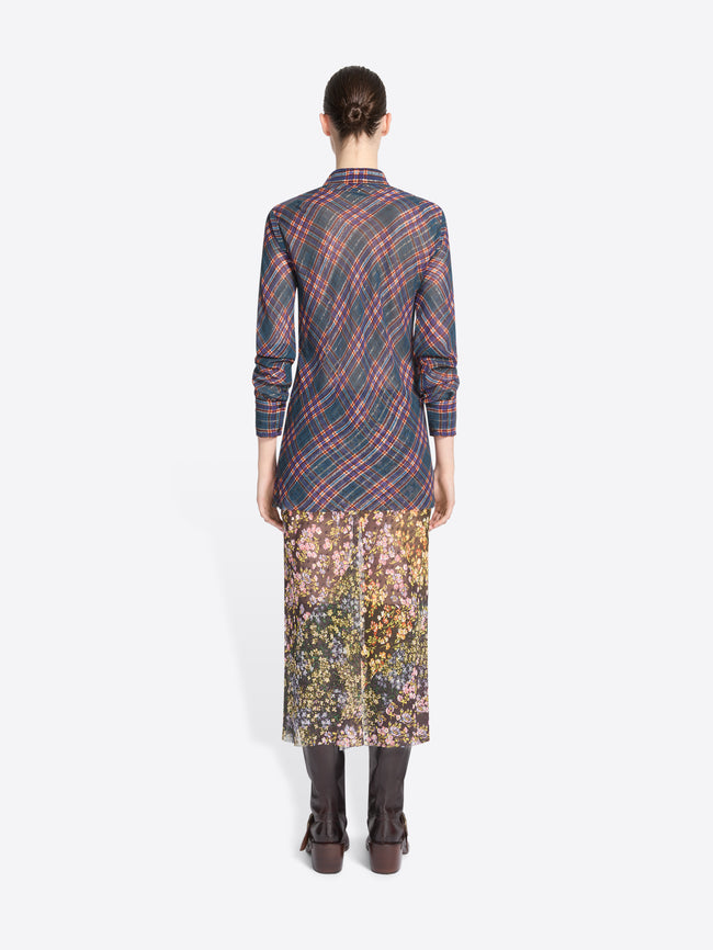 Back view of a model standing against a light gray background wearing a long-sleeve collared shirt with a muted plaid pattern in blue, red, and gray, paired with a floral midi skirt and dark brown knee-high boots, holding a small patterned handbag.