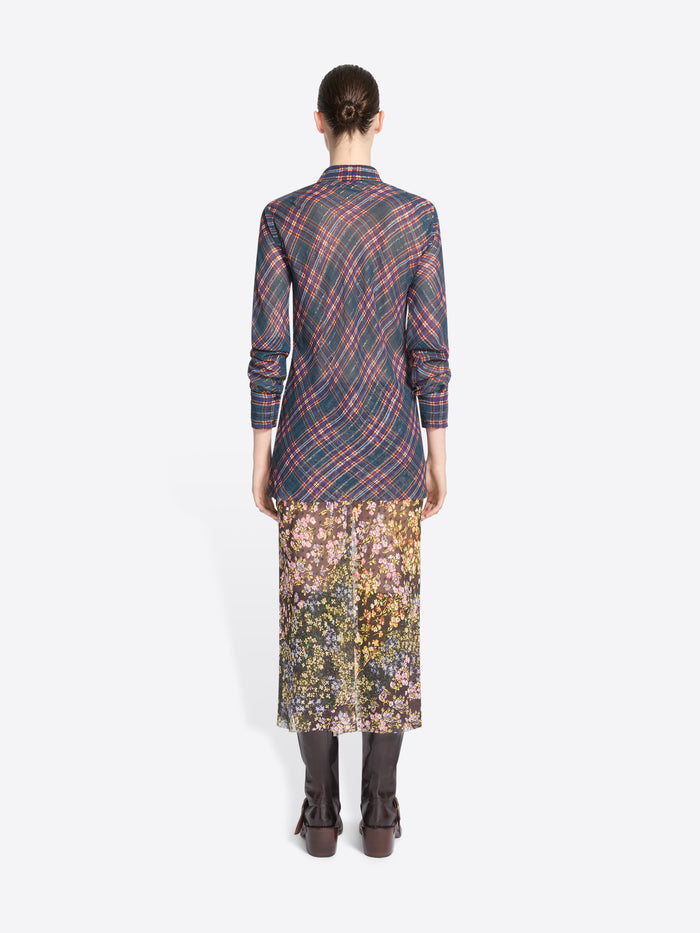 Back view of a model standing against a light gray background wearing a long-sleeve collared shirt with a muted plaid pattern in blue, red, and gray, paired with a floral midi skirt and dark brown knee-high boots, holding a small patterned handbag.