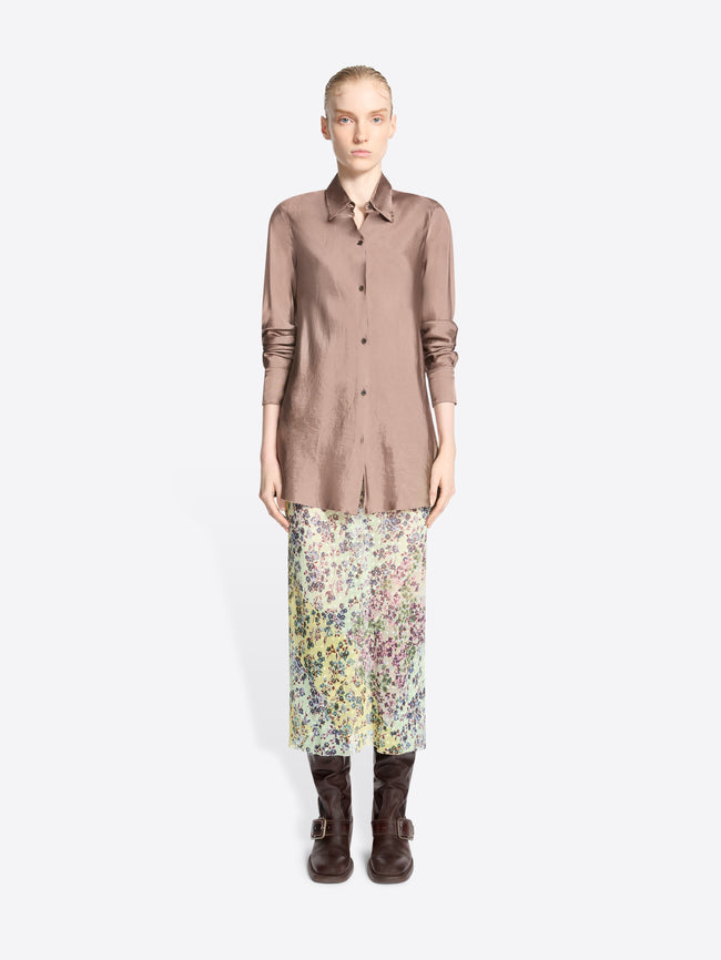 A model stands against a light gray background wearing a long-sleeve button-up shirt in a muted dusty rose, a multicolor floral midi skirt, and dark brown leather boots.