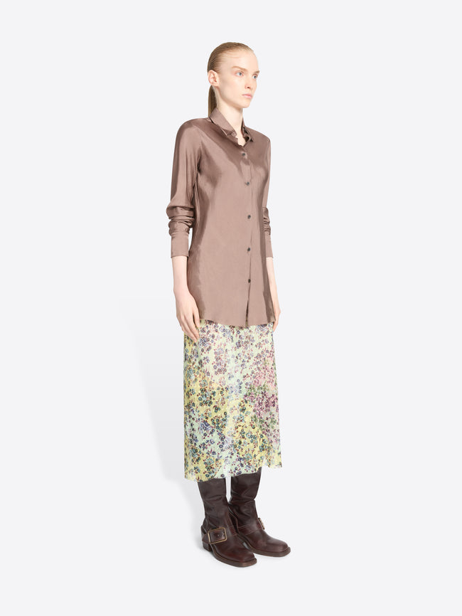 A model stands against a light gray background wearing a long-sleeve button-up shirt in a muted dusty rose, a multicolor floral midi skirt, and dark brown leather boots.