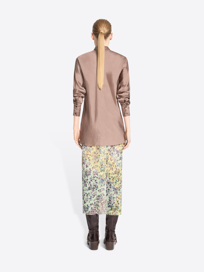 Back view of a model standing against a light gray background wearing a long-sleeve button-up shirt in a muted dusty rose, a multicolor floral midi skirt, and dark brown leather boots.