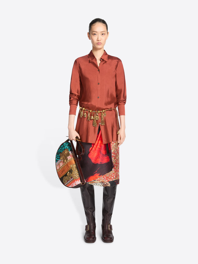 A model stands against a light gray background wearing a long-sleeve button-up shirt in a muted terracotta, a patterned skirt with red and black panels, a gold-toned belt with hanging charms, brown knee-high boots, and holding a colorful patterned handbag.