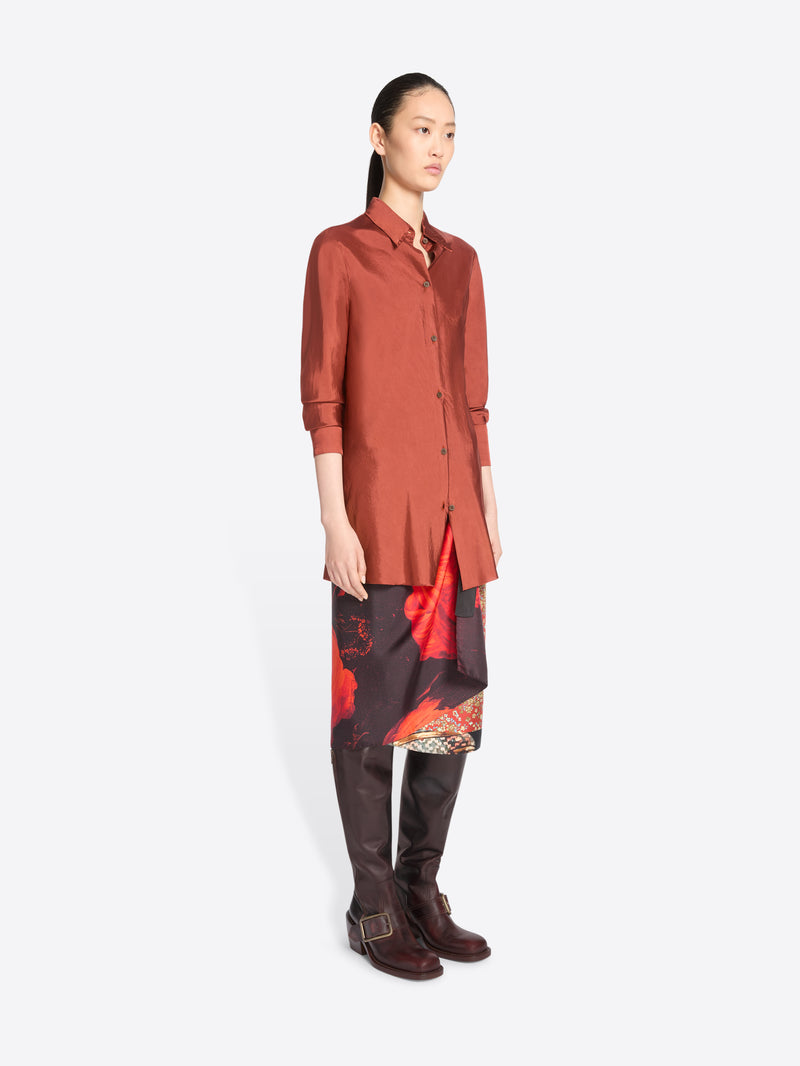A model stands against a light gray background wearing a long-sleeve button-up shirt in a muted terracotta, a patterned skirt with red and black panels and brown knee-high boots.
