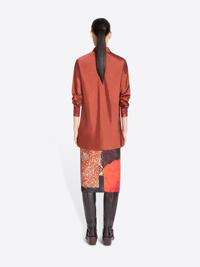 Back view of a model standing against a light gray background wearing a long-sleeve button-up shirt in a muted terracotta, a patterned skirt with red and black panels and brown knee-high boots.