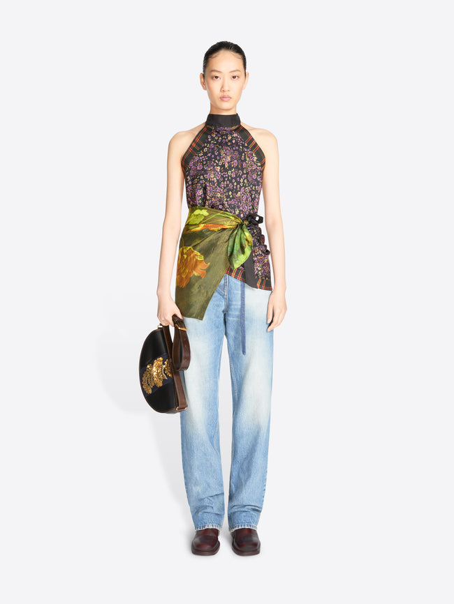 A model stands against a light gray background wearing a sleeveless high-neck top with a dark floral print and a green painterly wrap detail at the waist, paired with light blue jeans and holding a small dark handbag.