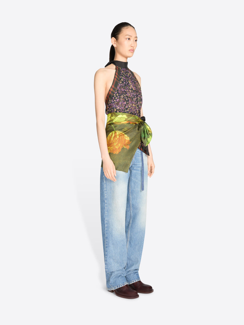 A model stands against a light gray background wearing a sleeveless high-neck top with a dark floral print and a green painterly wrap detail at the waist, paired with light blue jeans.