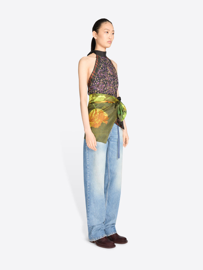 A model stands against a light gray background wearing a sleeveless high-neck top with a dark floral print and a green painterly wrap detail at the waist, paired with light blue jeans.