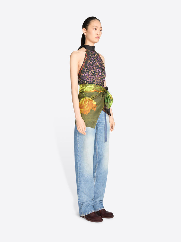 A model stands against a light gray background wearing a sleeveless high-neck top with a dark floral print and a green painterly wrap detail at the waist, paired with light blue jeans.