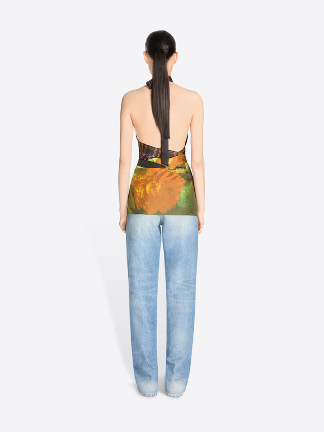Back view of a model standing against a light gray background wearing a sleeveless high-neck top with a dark floral print and a green painterly wrap detail at the waist, paired with light blue jeans.