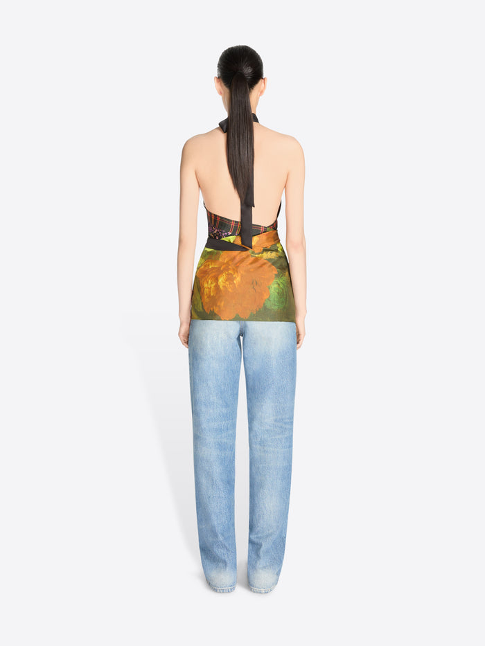 Back view of a model standing against a light gray background wearing a sleeveless high-neck top with a dark floral print and a green painterly wrap detail at the waist, paired with light blue jeans.