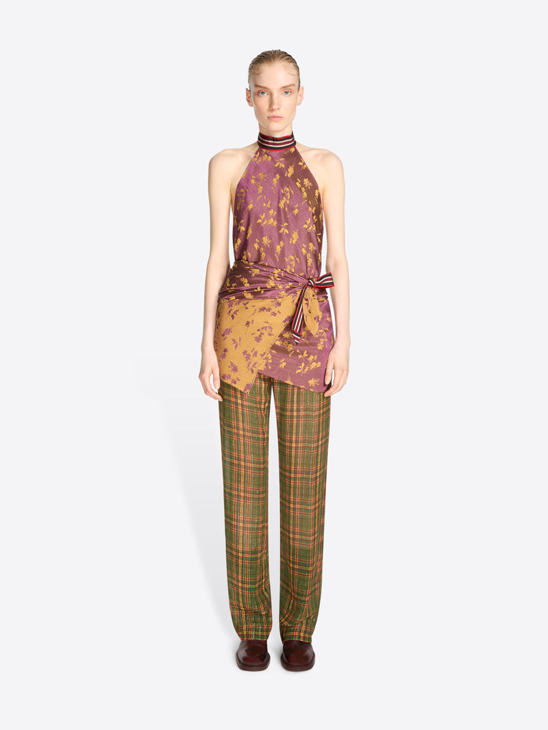 A model stands against a light gray background wearing a sleeveless halter top with a purple and gold abstract print, tied at the waist, paired with green and brown plaid trousers.