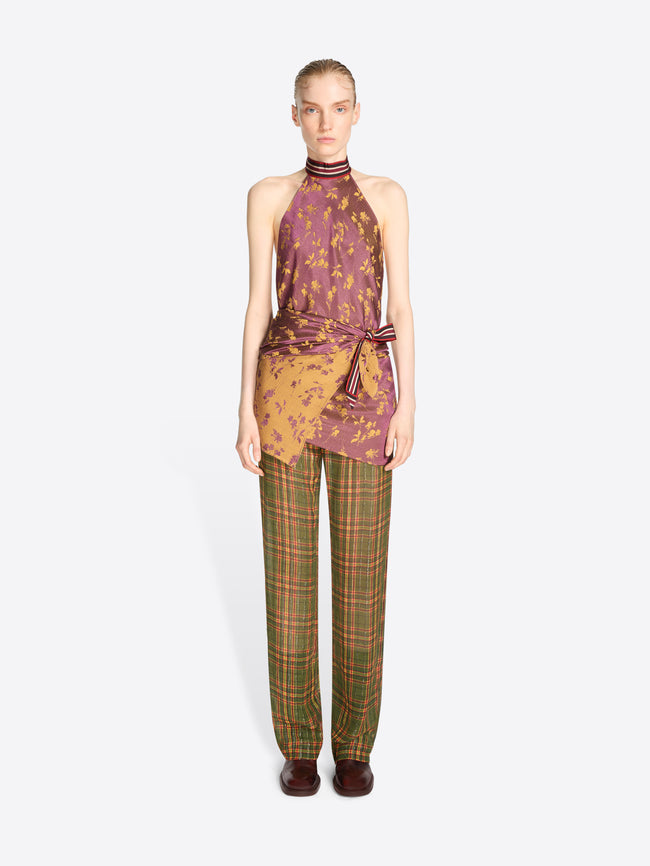 A model stands against a light gray background wearing a sleeveless halter top with a purple and gold abstract print, tied at the waist, paired with green and brown plaid trousers.