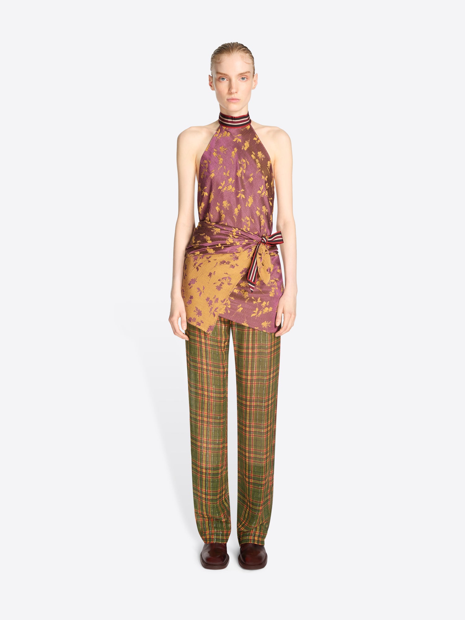 A model stands against a light gray background wearing a sleeveless halter top with a purple and gold abstract print, tied at the waist, paired with green and brown plaid trousers.