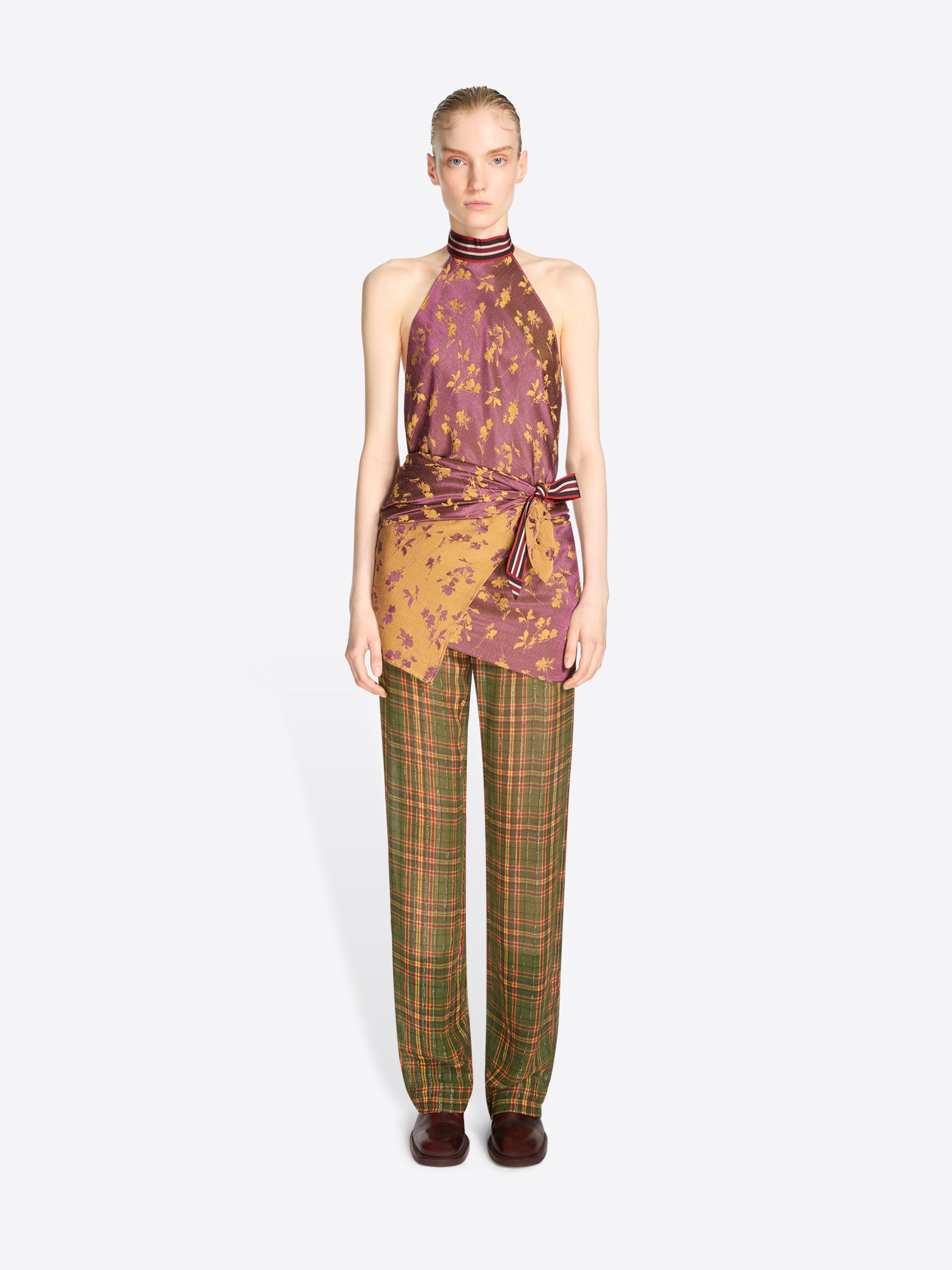 A model stands against a light gray background wearing a sleeveless halter top with a purple and gold abstract print, tied at the waist, paired with green and brown plaid trousers.