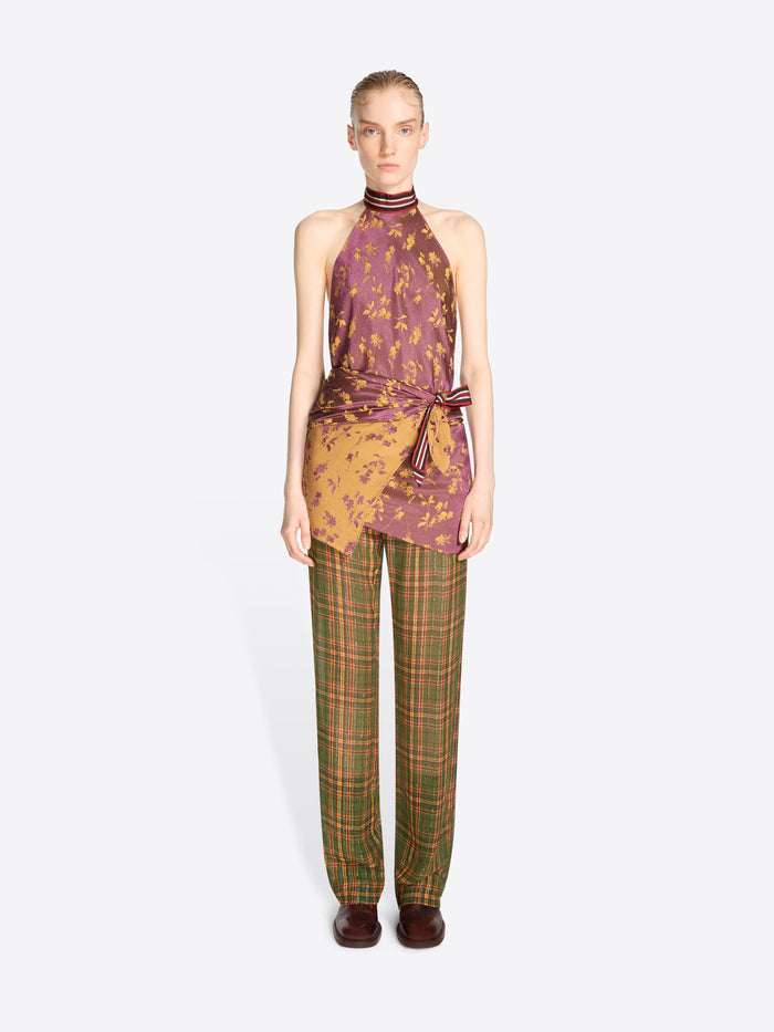 A model stands against a light gray background wearing a sleeveless halter top with a purple and gold abstract print, tied at the waist, paired with green and brown plaid trousers.