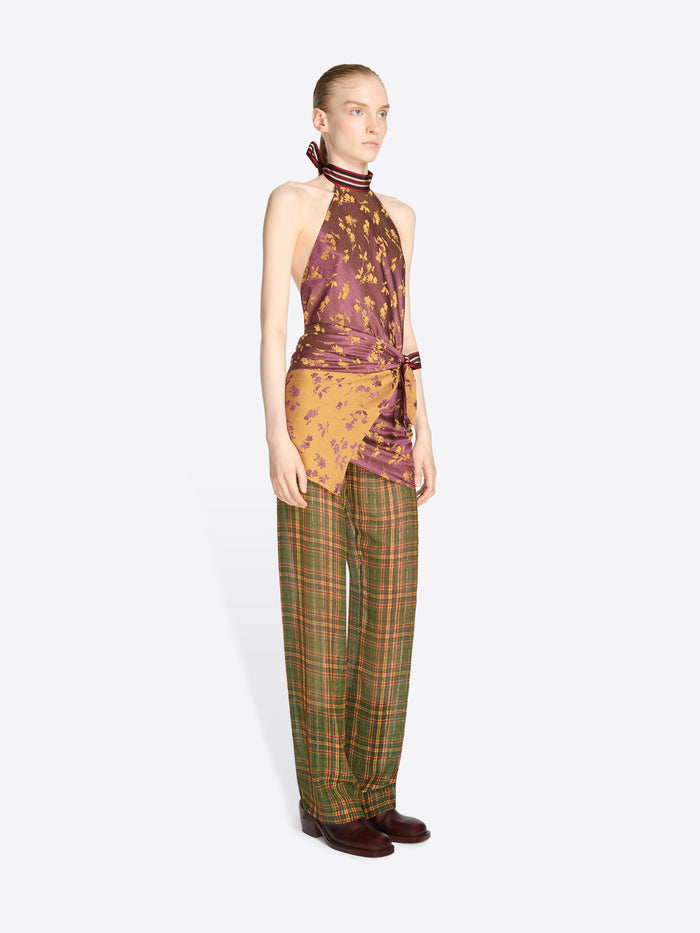 A model stands against a light gray background wearing a sleeveless halter top with a purple and gold abstract print, tied at the waist, paired with green and brown plaid trousers.