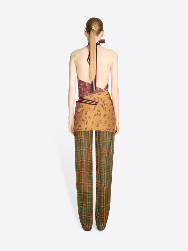 Back view of a model standing against a light gray background wearing a sleeveless halter top with a purple and gold abstract print, tied at the waist, paired with green and brown plaid trousers.