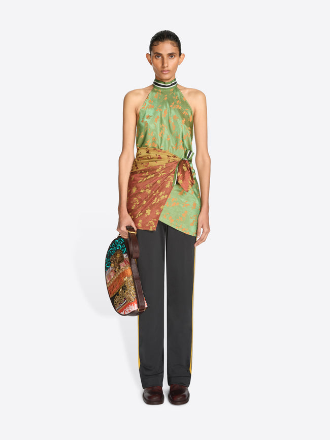 A model stands against a light gray background wearing a halter-neck wrap top with a green and gold patterned upper section and a rust-orange patterned lower section tied at the waist, paired with dark gray pants and holding a matching patterned handbag.