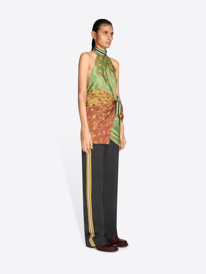 A model stands against a light gray background wearing a halter-neck wrap top with a green and gold patterned upper section and a rust-orange patterned lower section tied at the waist, paired with dark gray pants.