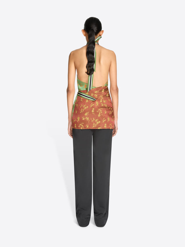 Back view of a model standing against a light gray background wearing a halter-neck wrap top with a green and gold patterned upper section and a rust-orange patterned lower section tied at the waist, paired with dark gray pants.