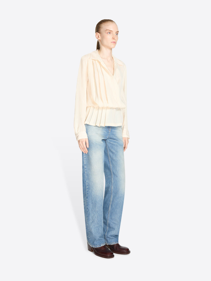 A model stands against a light gray background wearing a cream long-sleeve ruffled blouse, light blue jeans and dark brown shoes.