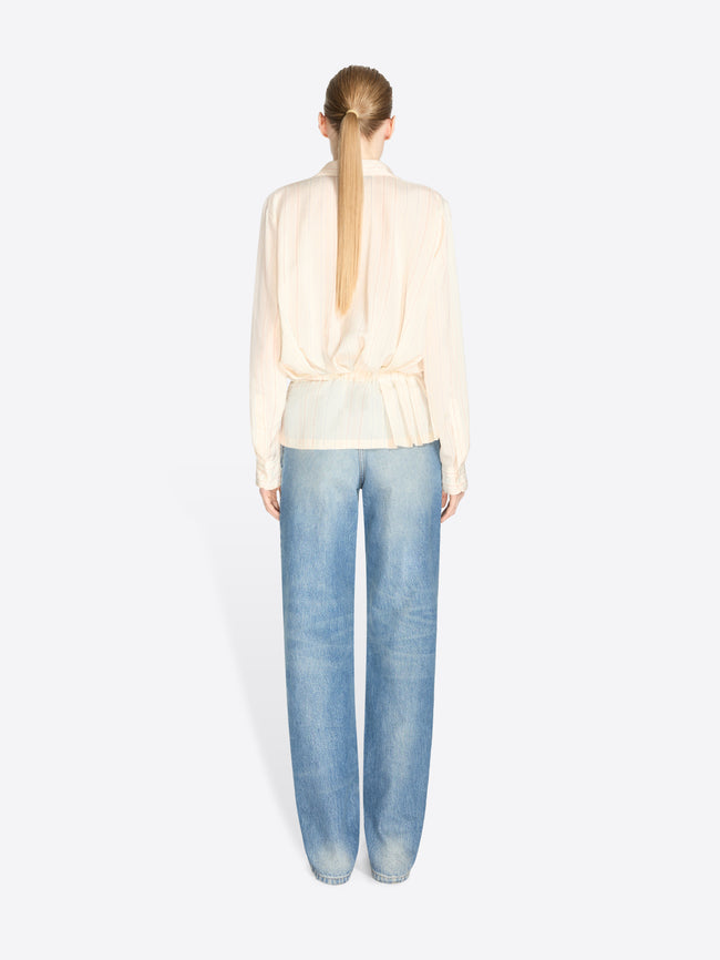 Back view of a model standing against a light gray background wearing a cream long-sleeve ruffled blouse, light blue jeans and dark brown shoes.