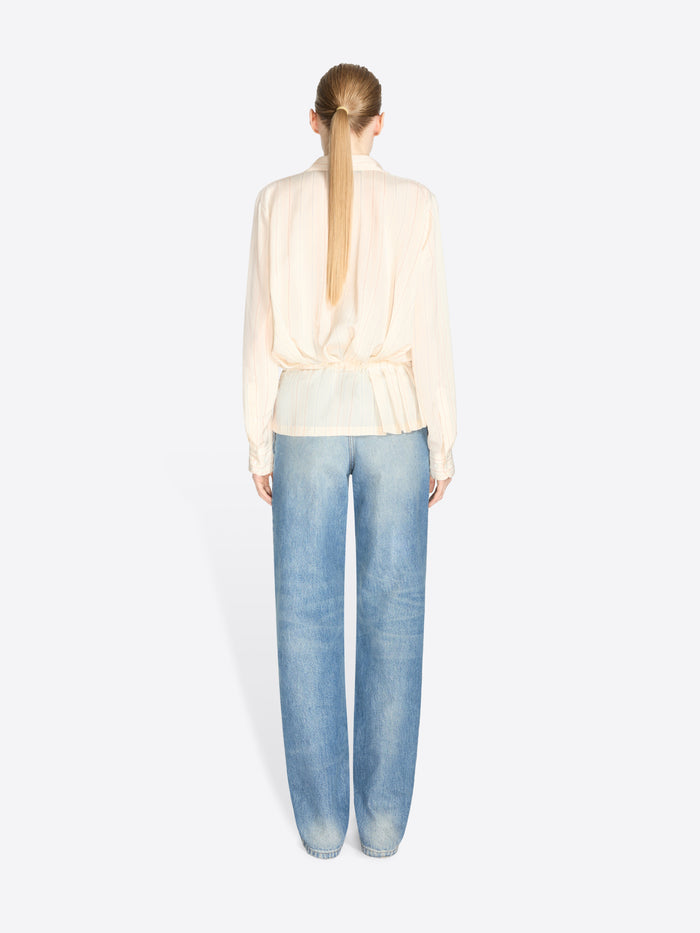Back view of a model standing against a light gray background wearing a cream long-sleeve ruffled blouse, light blue jeans and dark brown shoes.