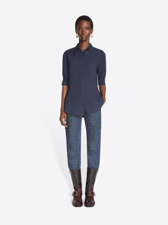 A model stands against a light gray background wearing a navy blue button-up shirt with three-quarter sleeves, blue plaid pants, and dark brown leather boots.