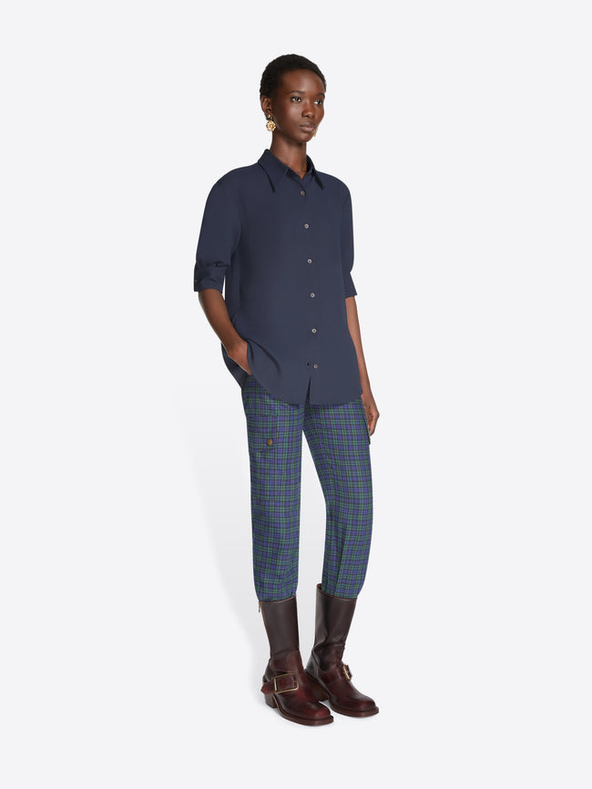 A model stands against a light gray background wearing a navy blue button-up shirt with three-quarter sleeves, blue plaid pants, and dark brown leather boots.