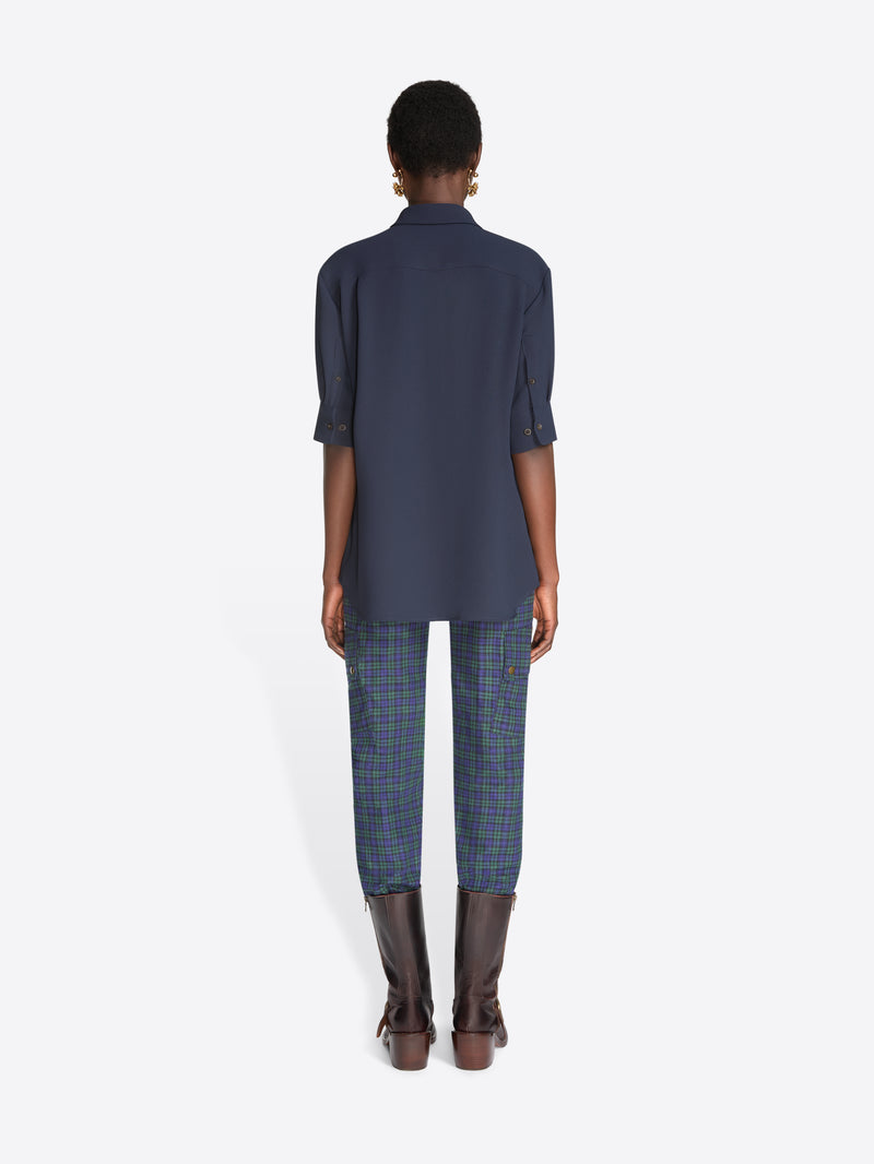 Back view of a model standing against a light gray background wearing a navy blue button-up shirt with three-quarter sleeves, blue plaid pants, and dark brown leather boots.