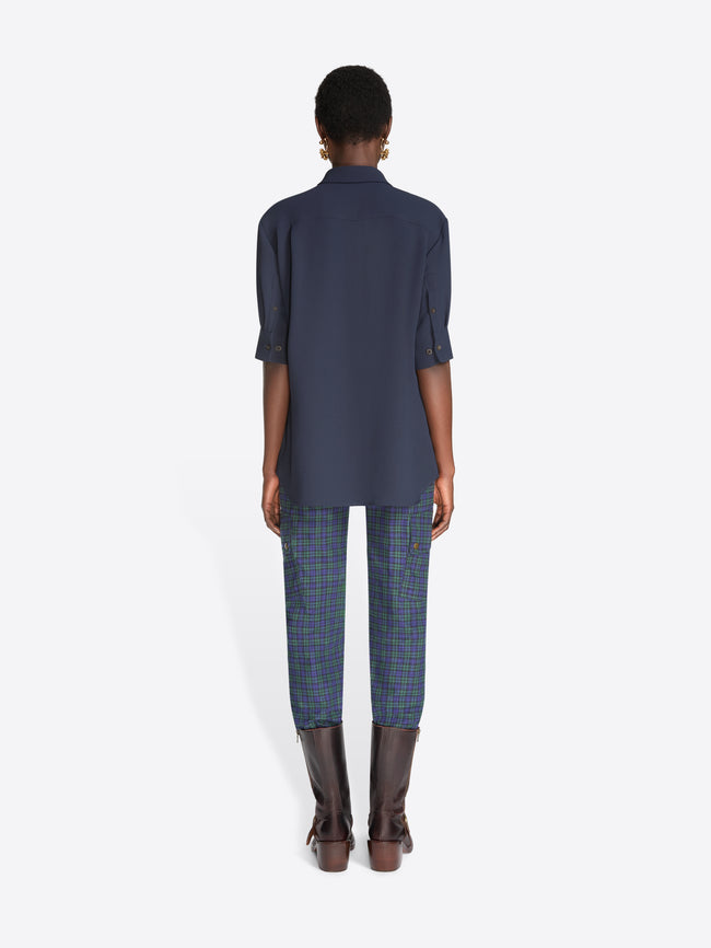 Back view of a model standing against a light gray background wearing a navy blue button-up shirt with three-quarter sleeves, blue plaid pants, and dark brown leather boots.