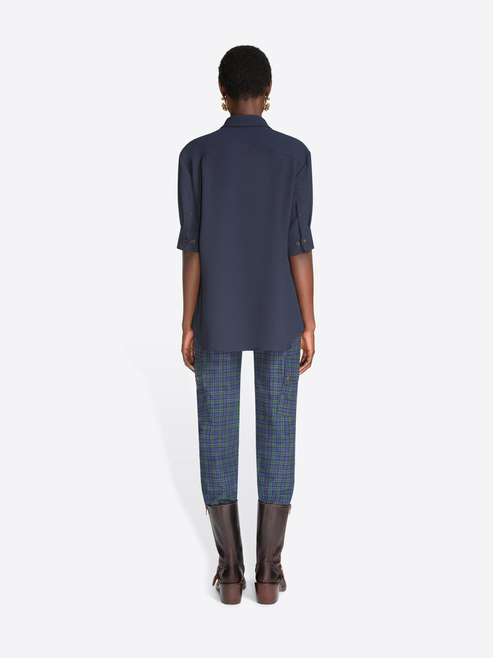 Back view of a model standing against a light gray background wearing a navy blue button-up shirt with three-quarter sleeves, blue plaid pants, and dark brown leather boots.